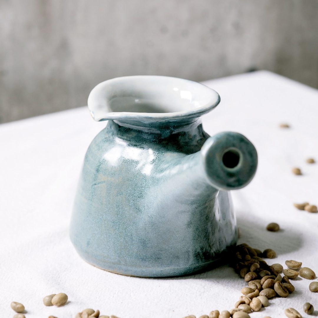 Blue craft ceramic coffee pot Turkish cezve standing on white tablecloth with coffee beans above. Gray wall behind. Copy space.